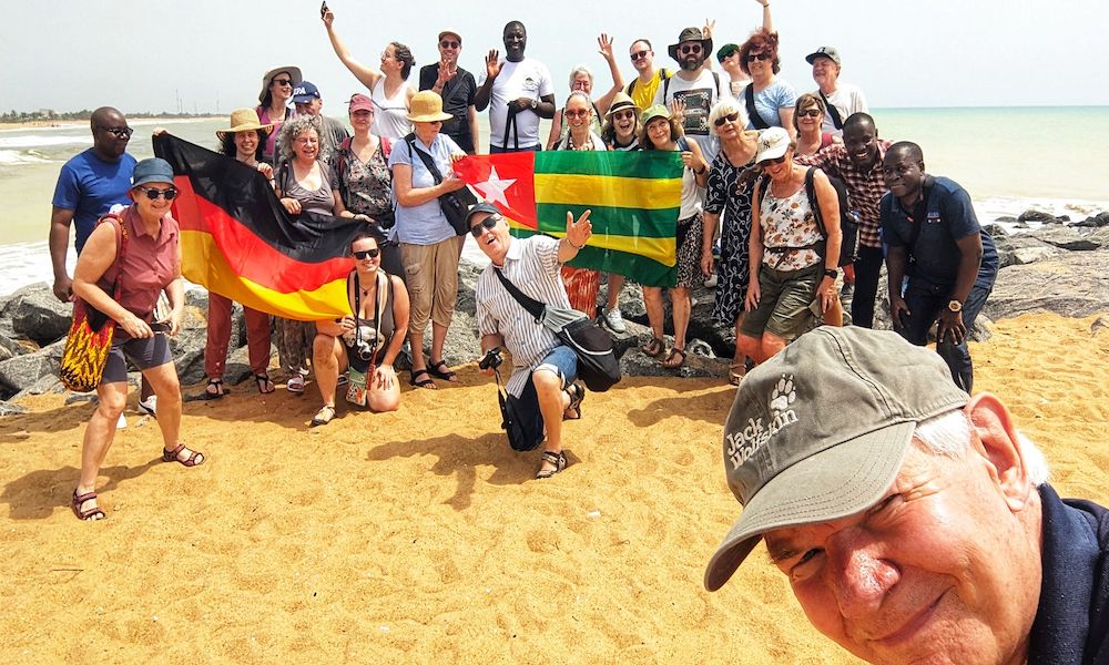 Eine fröhliche Gruppe von Menschen steht am Strand vor dem Meer und posiert für ein Gruppenfoto. Einige halten die Flaggen von Deutschland und Togo.  Im Vordergrund macht ein älterer Mann mit grauer Kappe ein Selfie mit der Gruppe im Hintergrund. Der Himmel ist hell, das Wetter sonnig.